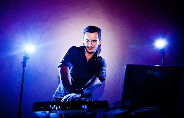 DJ im Spotlicht am Pult, dunkle Bühne, Clubatmosphäre.