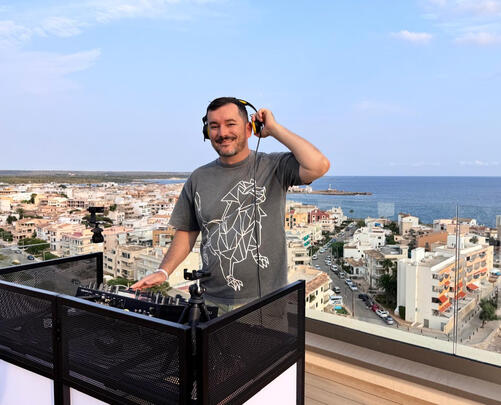 DJ auf Dachterrasse mit Skyline im Hintergrund, Kopfhörer am Ohr.