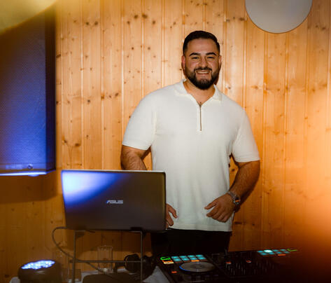 DJ lächelt am Pult vor Holz-Wand, warmes Licht.
